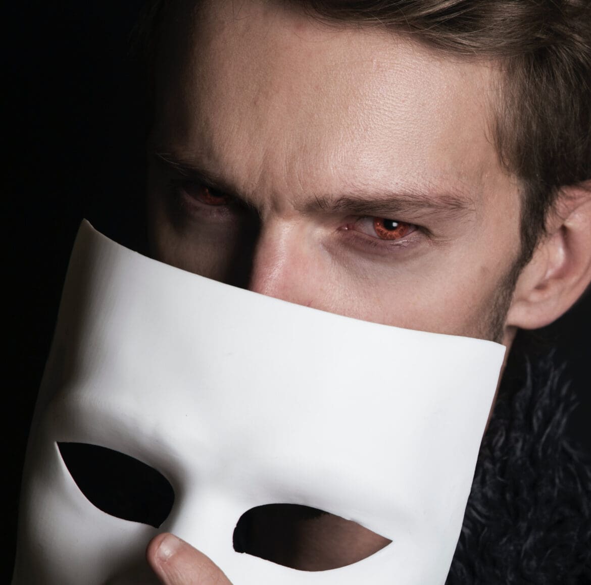 Vampire a man with red eyes holding a white mask over his face